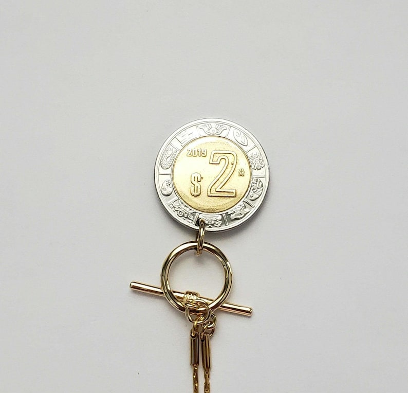 K&ouml;nnte beinhalten: Ein zweifarbiger M&uuml;nzanh&auml;nger mit einem silbernen Au&szlig;enring und einem goldenen Zentrum, das den Text $2 und 2019 tr&auml;gt. Die M&uuml;nze ist an einem goldfarbenen Knebelverschluss mit einem runden Ring und einem Querstab befestigt, der an einer feinen Goldkette h&auml;ngt.