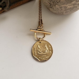 May include: A gold necklace with a Singaporean coin pendant. The coin features the Singapore coat of arms and the year 1995. The necklace has a toggle clasp.