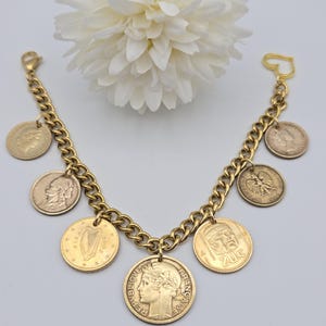 May include: A gold-toned chain link charm bracelet featuring various international coins as charms. The coins include depictions of a harp, a profile of a person, and the text Republique Francaise. The bracelet has a heart-shaped clasp closure.