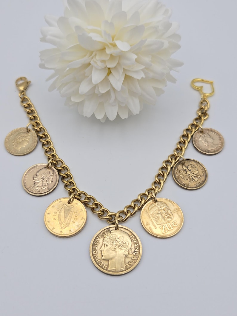 May include: A gold-toned chain link charm bracelet featuring various international coins as charms. The coins include depictions of a harp, a profile of a person, and the text Republique Francaise. The bracelet has a heart-shaped clasp closure.