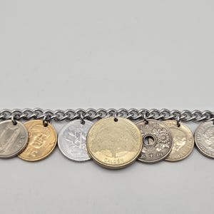 Pode incluir: Uma pulseira de corrente de metal prateado com uma s&eacute;rie de moedas internacionais penduradas. As moedas variam na cor, incluindo tons de ouro e prata, e apresentam diversos desenhos como uma &aacute;rvore, uma harpa e motivos florais com texto.
