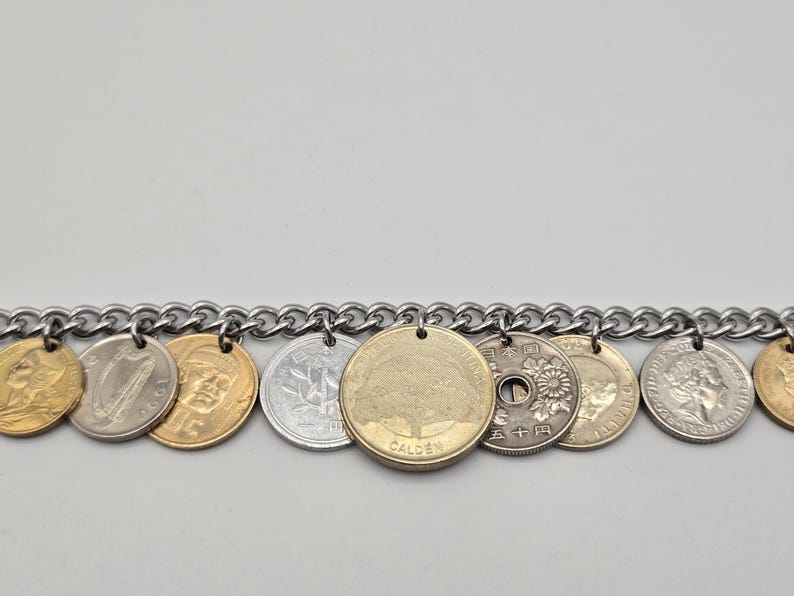 Pode incluir: Uma pulseira de corrente de metal prateado com uma s&eacute;rie de moedas internacionais penduradas. As moedas variam na cor, incluindo tons de ouro e prata, e apresentam diversos desenhos como uma &aacute;rvore, uma harpa e motivos florais com texto.