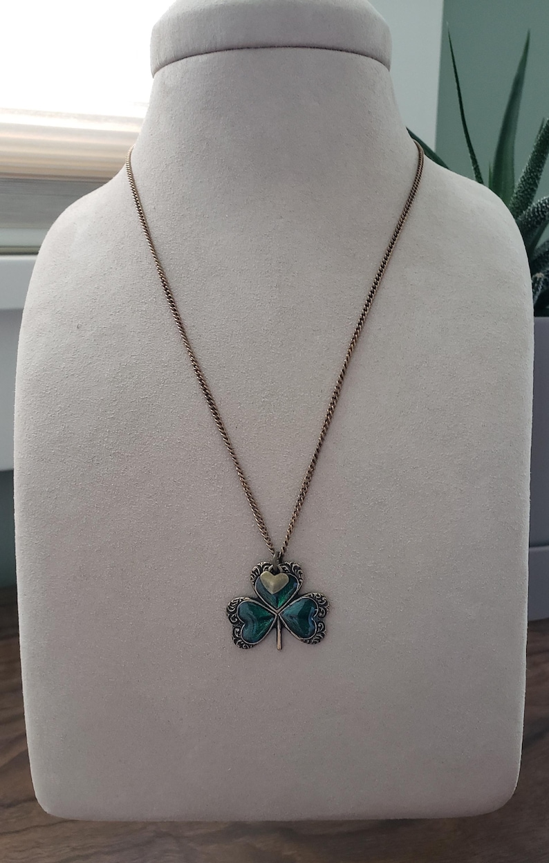 May include: A gold chain necklace with a green shamrock pendant. The shamrock is made of three heart-shaped leaves with a gold heart in the centre.