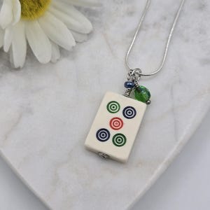 May include: A silver-toned snake chain necklace featuring a rectangular off-white mahjong tile pendant. The tile displays five circular bullseye patterns in green, blue, and red. A small blue bead and a translucent green bead hang above the tile.