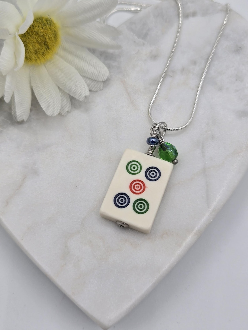 May include: A silver-toned snake chain necklace featuring a rectangular off-white mahjong tile pendant. The tile displays five circular bullseye patterns in green, blue, and red. A small blue bead and a translucent green bead hang above the tile.