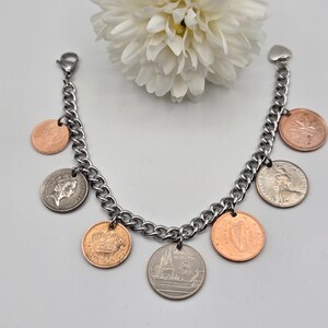 May include: A silver-toned metal chain charm bracelet featuring seven assorted international coins as pendants. The coins vary in color between copper and silver tones, displaying various national symbols, portraits, and text. A heart-shaped clasp is attached.