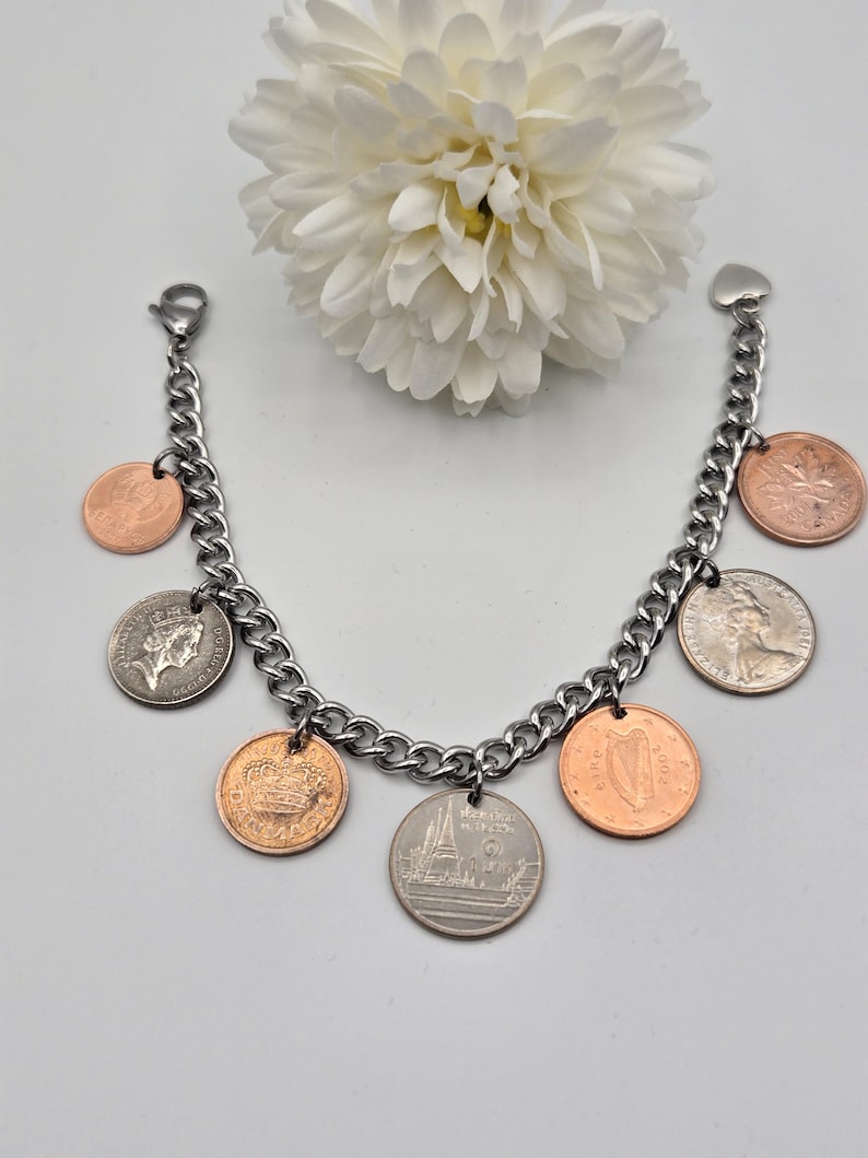May include: A silver-toned metal chain charm bracelet featuring seven assorted international coins as pendants. The coins vary in color between copper and silver tones, displaying various national symbols, portraits, and text. A heart-shaped clasp is attached.