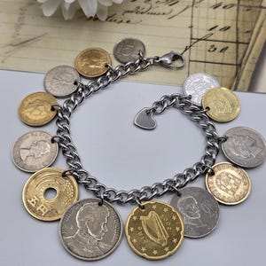 Pode incluir: Uma pulseira de elos de metal prateado com uma cole&ccedil;&atilde;o de v&aacute;rias moedas internacionais como berloques. As moedas variam em tamanho, cor e acabamento met&aacute;lico, incluindo tons dourados e prateados. Um pequeno berloque em forma de cora&ccedil;&atilde;o est&aacute; preso.