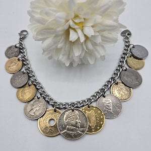 Pode incluir: Uma pulseira de berloques com uma corrente de metal prateado adornada com v&aacute;rias moedas internacionais em tons de dourado e prateado. As moedas exibem diversos retratos, s&iacute;mbolos e textos, incluindo 10 CENT, 20, 2002 e REPUBLICA DE CHILE.