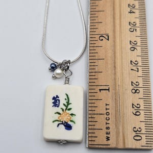 May include: A silver-toned snake chain necklace featuring a rectangular off-white tile pendant with a painted yellow flower and blue character. A small white pearl and a dark blue bead are attached to the bail. A ruler shows the pendant is 2.5 cm long.