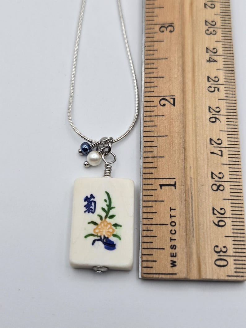May include: A silver-toned snake chain necklace featuring a rectangular off-white tile pendant with a painted yellow flower and blue character. A small white pearl and a dark blue bead are attached to the bail. A ruler shows the pendant is 2.5 cm long.