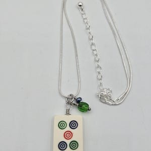 May include: A silver-toned snake chain necklace featuring a rectangular off-white mahjong tile pendant. The tile displays five circular symbols in green, blue, and red. A small green faceted bead and a dark blue bead hang beside the tile.