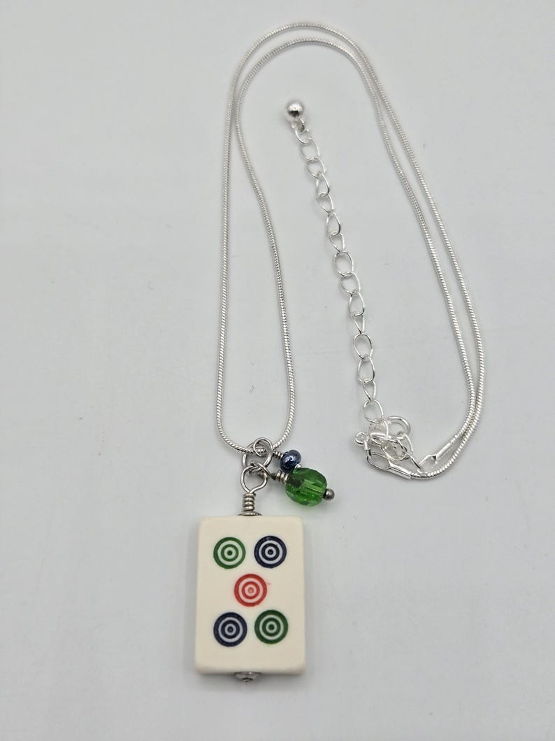 May include: A silver-toned snake chain necklace featuring a rectangular off-white mahjong tile pendant. The tile displays five circular symbols in green, blue, and red. A small green faceted bead and a dark blue bead hang beside the tile.