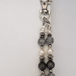 May include: A silver-toned keychain with a lobster clasp and a decorative beaded charm. The charm features a heart-shaped accent, alternating pearl and rhinestone-studded beads, and dark gray beads. The keychain is attached to a white spiral notebook.