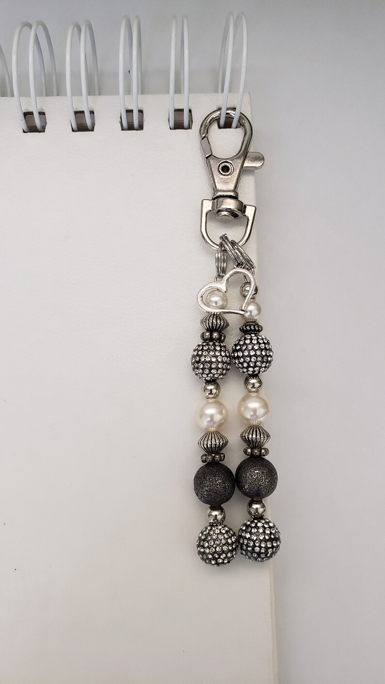 May include: A silver-toned keychain with a lobster clasp and a decorative beaded charm. The charm features a heart-shaped accent, alternating pearl and rhinestone-studded beads, and dark gray beads. The keychain is attached to a white spiral notebook.