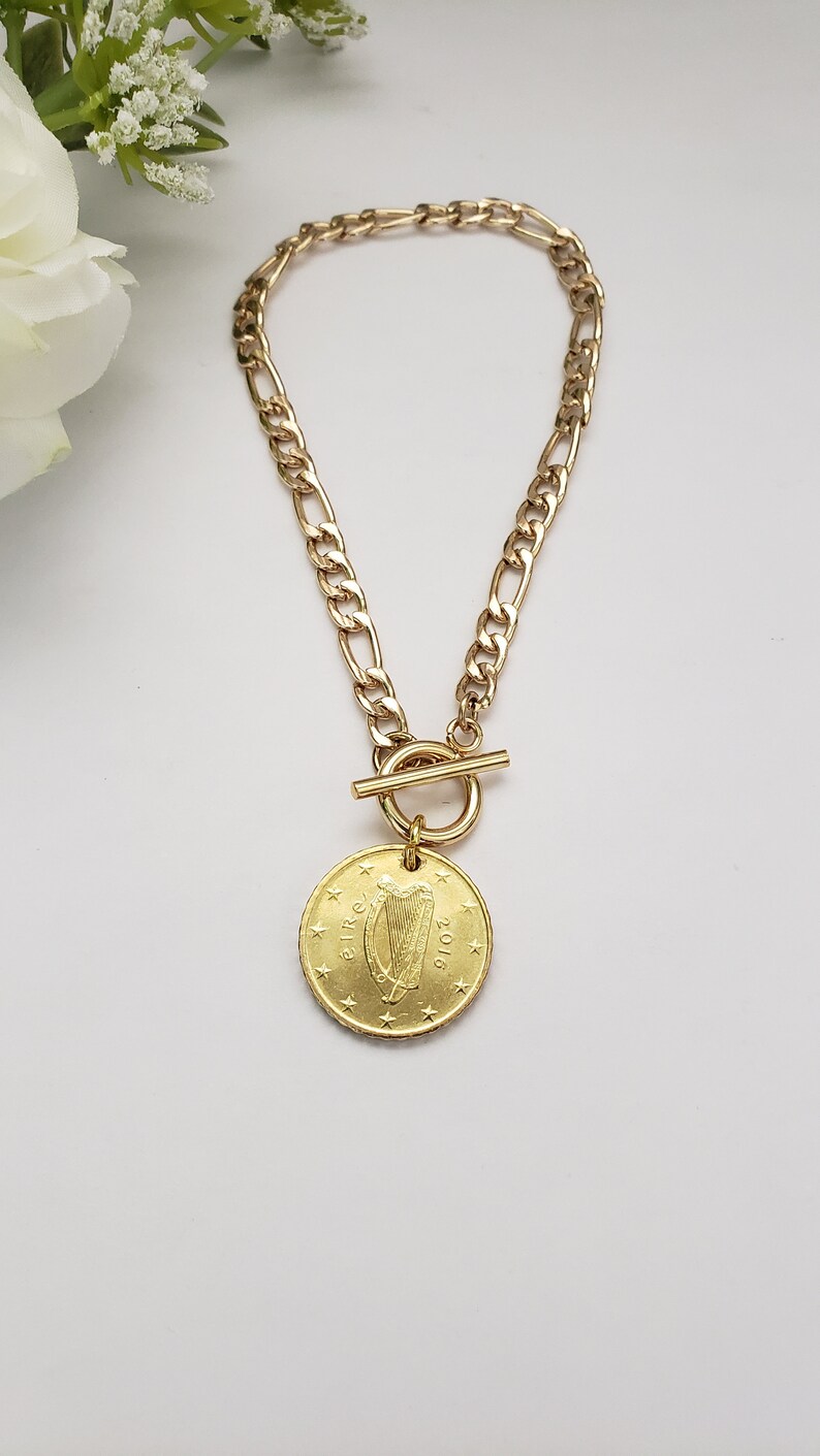 May include: A gold chain necklace with a toggle clasp and a gold coin pendant featuring the Irish harp and the year 2016.