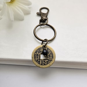 May include: Antique bronze keychain with a round coin pendant featuring Chinese characters and a small black bead. The keychain has a lobster clasp and a split ring for attaching keys. A decorative accessory.