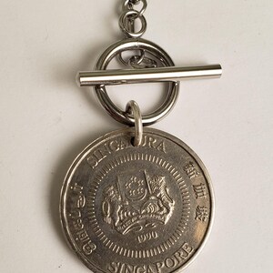 May include: A silver necklace with a toggle clasp and a Singapore dollar coin pendant featuring the national coat of arms and the year 1990.
