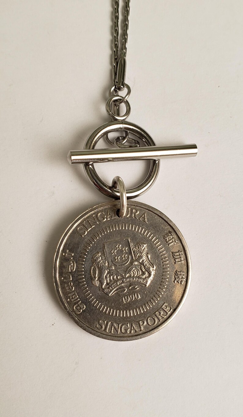 May include: A silver necklace with a toggle clasp and a Singapore dollar coin pendant featuring the national coat of arms and the year 1990.