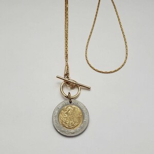 K&ouml;nnte beinhalten: Eine goldfarbene Kettenhalskette mit Knebelverschluss und einem runden Anh&auml;nger. Der Anh&auml;nger ist eine bimetallische M&uuml;nze mit einem goldfarbenen Zentrum, das einen Adler zeigt, und einem silberfarbenen Au&szlig;enring mit dem Text Estados Unidos Mexicanos.