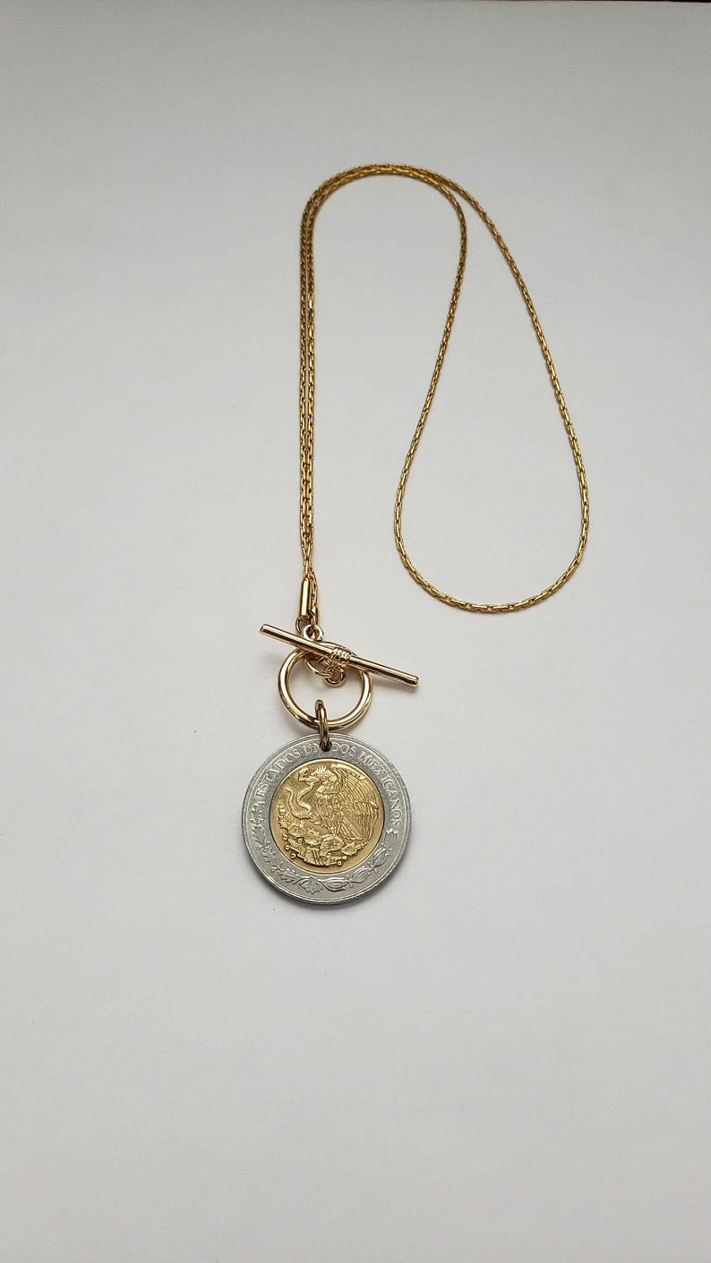 K&ouml;nnte beinhalten: Eine goldfarbene Kettenhalskette mit Knebelverschluss und einem runden Anh&auml;nger. Der Anh&auml;nger ist eine bimetallische M&uuml;nze mit einem goldfarbenen Zentrum, das einen Adler zeigt, und einem silberfarbenen Au&szlig;enring mit dem Text Estados Unidos Mexicanos.