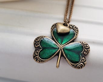 Puede incluir: Un collar de cadena dorada con un colgante de trébol verde. El trébol tiene un pequeño amuleto de corazón dorado en la parte superior.