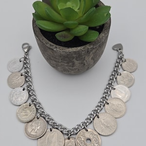 K&ouml;nnte beinhalten: Ein silbernes Charm-Armband mit einer klobigen Kette, verziert mit verschiedenen M&uuml;nzanh&auml;ngern. Das Armband hat eine Vielzahl von runden M&uuml;nzen in verschiedenen Gr&ouml;&szlig;en und Designs. Eine kleine, gr&uuml;ne Sukkulentenpflanze in einem grauen Topf befindet sich im Hintergrund.