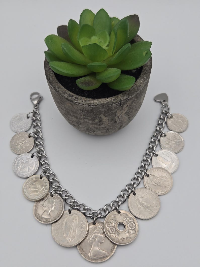 K&ouml;nnte beinhalten: Ein silbernes Charm-Armband mit einer klobigen Kette, verziert mit verschiedenen M&uuml;nzanh&auml;ngern. Das Armband hat eine Vielzahl von runden M&uuml;nzen in verschiedenen Gr&ouml;&szlig;en und Designs. Eine kleine, gr&uuml;ne Sukkulentenpflanze in einem grauen Topf befindet sich im Hintergrund.