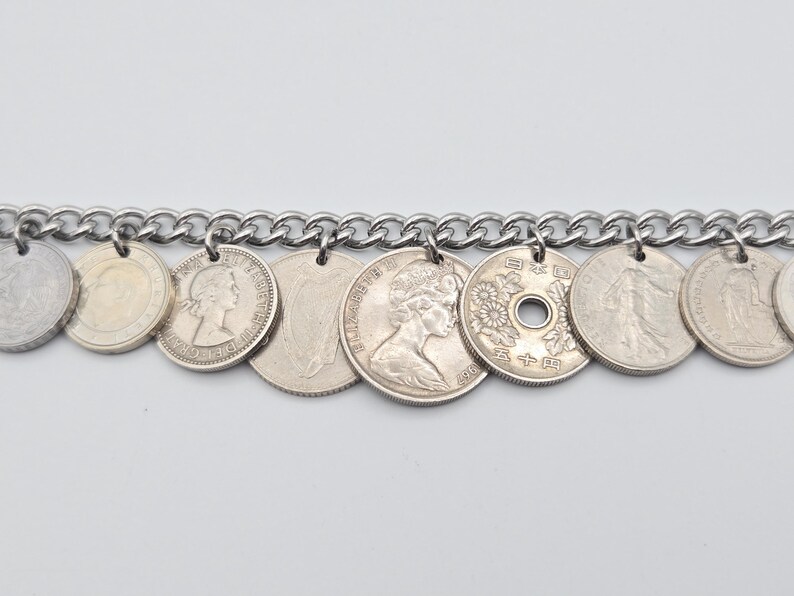 K&ouml;nnte beinhalten: Ein silbernes Kettenarmband mit verschiedenen Vintage-M&uuml;nzen als Anh&auml;nger. Die M&uuml;nzen, in verschiedenen Gr&ouml;&szlig;en und Designs, sind mit kleinen Schlaufen an der Kette befestigt. Das Armband wird vor einem schlichten wei&szlig;en Hintergrund pr&auml;sentiert.