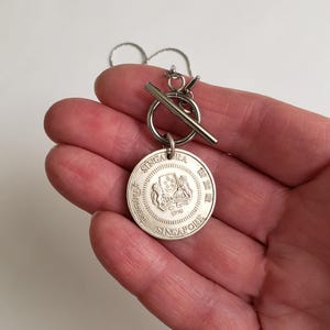 May include: Silver necklace with a round pendant featuring the Singapore coat of arms and the text "Singapore" and "1990". The necklace has a silver toggle clasp.