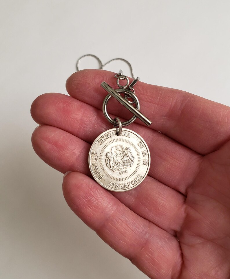 May include: Silver necklace with a round pendant featuring the Singapore coat of arms and the text "Singapore" and "1990". The necklace has a silver toggle clasp.