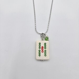May include: A silver-toned chain necklace featuring a rectangular off-white mahjong tile pendant. The tile displays a central red symbol flanked by two vertical green symbols. A small white pearl and a green crystal bead accent the top jump ring.