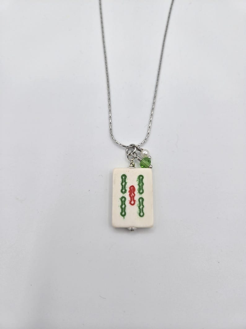 May include: A silver-toned chain necklace featuring a rectangular off-white mahjong tile pendant. The tile displays a central red symbol flanked by two vertical green symbols. A small white pearl and a green crystal bead accent the top jump ring.