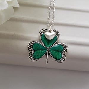 May include: A silver necklace featuring a green shamrock pendant with a small silver heart charm. The shamrock has an intricate design and is accented with green enamel.