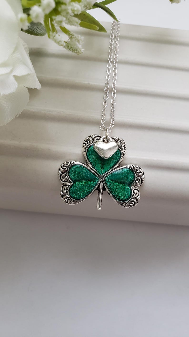 May include: A silver necklace featuring a green shamrock pendant with a small silver heart charm. The shamrock has an intricate design and is accented with green enamel.