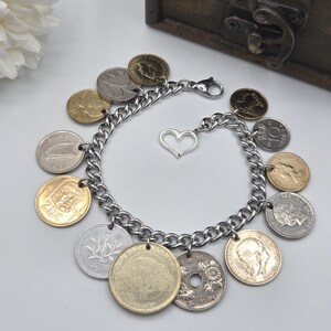 Pode incluir: Uma pulseira de elos em tom prateado com v&aacute;rias moedas internacionais como berloques. A pulseira inclui um pequeno berloque em forma de cora&ccedil;&atilde;o prateado e um fecho lagosta. As moedas variam na cor, incluindo tons de ouro, prata e bronze, e apresentam diversos retratos, s&iacute;mbolos e textos.