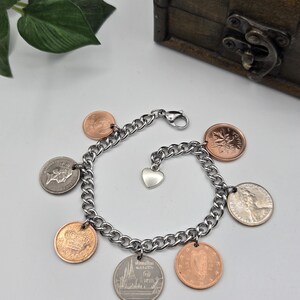 May include: A silver-toned metal chain link charm bracelet featuring seven assorted international coins in copper and silver colors, including a Canadian maple leaf coin, a Thai coin, and a small silver heart-shaped charm attached to the clasp.