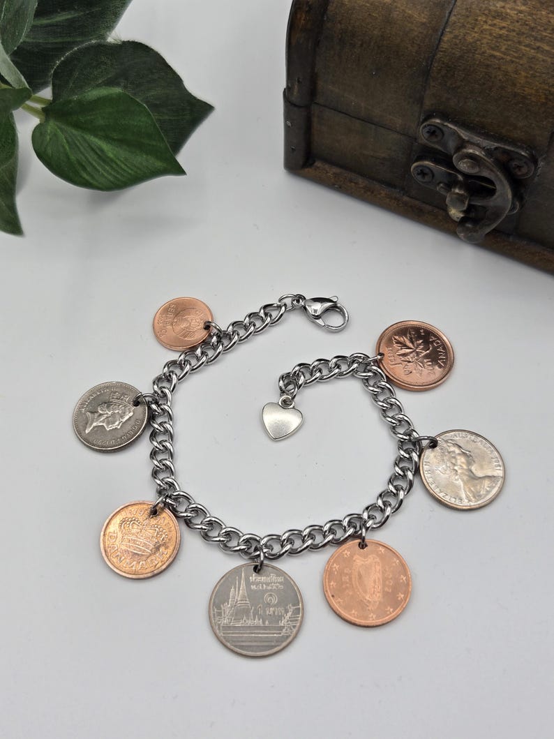 May include: A silver-toned metal chain link charm bracelet featuring seven assorted international coins in copper and silver colors, including a Canadian maple leaf coin, a Thai coin, and a small silver heart-shaped charm attached to the clasp.