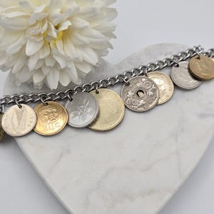 Pode incluir: Uma pulseira de berloques com uma corrente de metal prateado adornada com v&aacute;rias moedas internacionais em tons de dourado e prateado. As moedas exibem designs diversos, incluindo uma harpa, um rosto, padr&otilde;es florais e texto, penduradas na corrente.