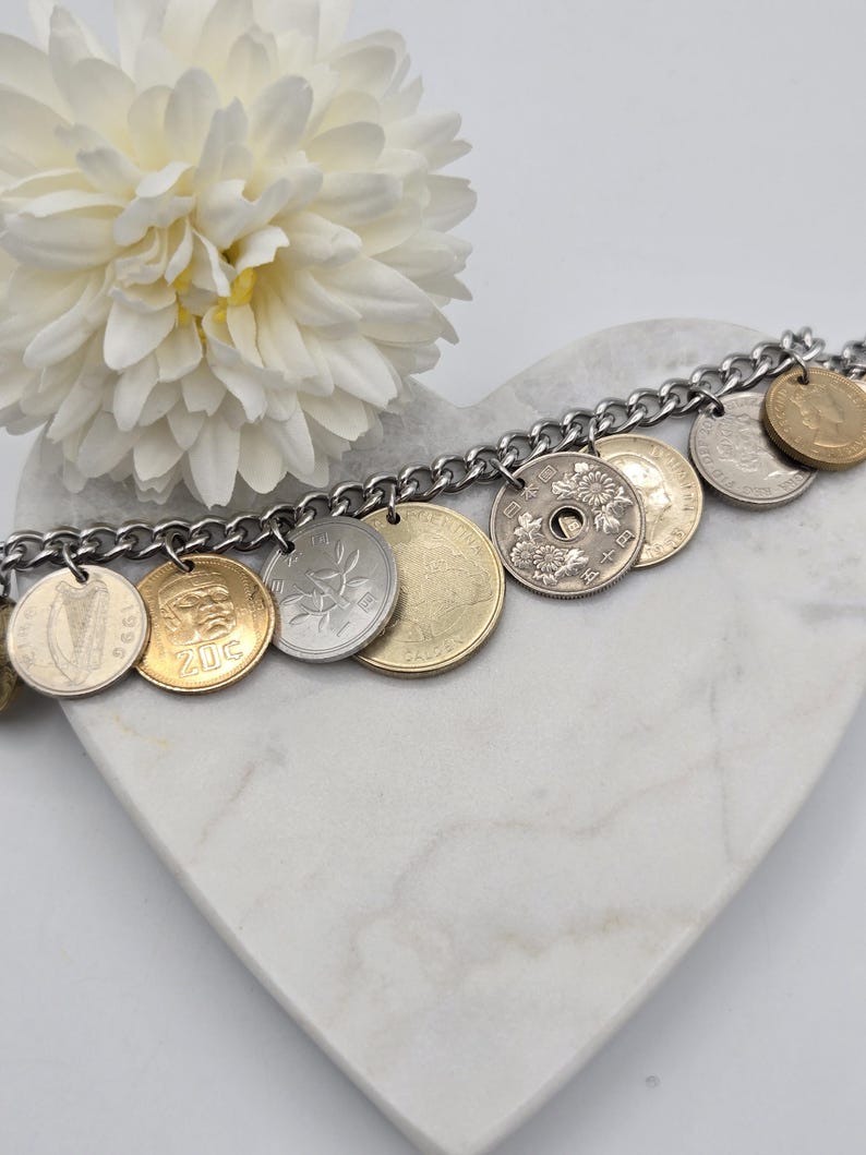 Pode incluir: Uma pulseira de berloques com uma corrente de metal prateado adornada com v&aacute;rias moedas internacionais em tons de dourado e prateado. As moedas exibem designs diversos, incluindo uma harpa, um rosto, padr&otilde;es florais e texto, penduradas na corrente.