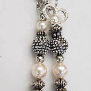 May include: A silver-toned keychain with a lobster clasp and multiple beaded strands. The strands feature faux pearls, rhinestone-studded spheres, and textured dark gray beads, with a heart-shaped charm.
