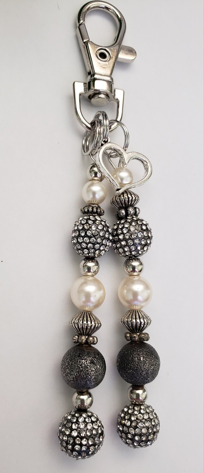 May include: A silver-toned keychain with a lobster clasp and multiple beaded strands. The strands feature faux pearls, rhinestone-studded spheres, and textured dark gray beads, with a heart-shaped charm.
