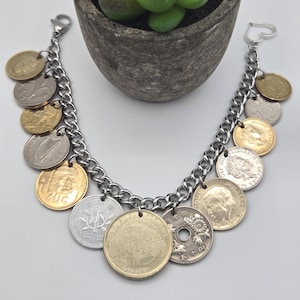 Pode incluir: Uma pulseira de corrente de metal prateado com uma cole&ccedil;&atilde;o de v&aacute;rias moedas internacionais como pingentes. As moedas variam em cor, incluindo tons de ouro, prata e bronze, e apresentam diversos desenhos, retratos e textos de v&aacute;rios pa&iacute;ses.