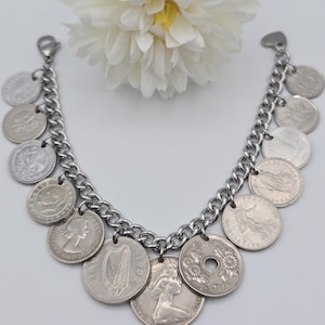 K&ouml;nnte beinhalten: Silberfarbenes Charm-Armband mit Kettendesign und verschiedenen M&uuml;nzanh&auml;ngern. Die M&uuml;nzen sind unterschiedlich gro&szlig; und zeigen Portr&auml;ts und Symbole. Das Armband wird vor einem wei&szlig;en Hintergrund mit einer wei&szlig;en Blume pr&auml;sentiert.