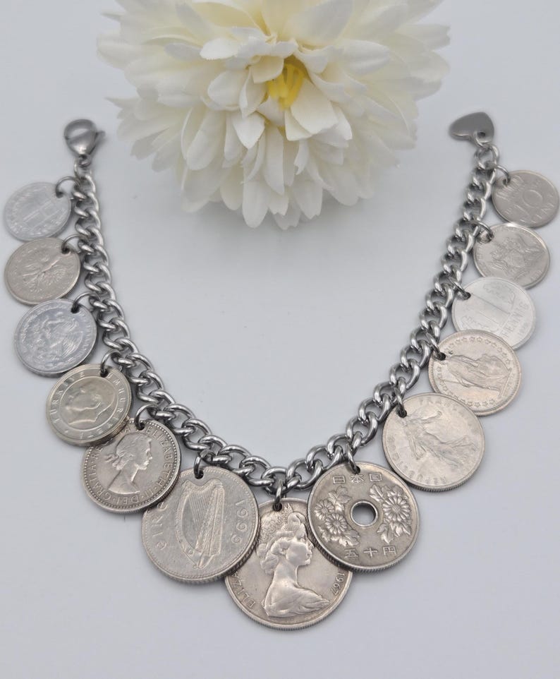 K&ouml;nnte beinhalten: Silberfarbenes Charm-Armband mit Kettendesign und verschiedenen M&uuml;nzanh&auml;ngern. Die M&uuml;nzen sind unterschiedlich gro&szlig; und zeigen Portr&auml;ts und Symbole. Das Armband wird vor einem wei&szlig;en Hintergrund mit einer wei&szlig;en Blume pr&auml;sentiert.