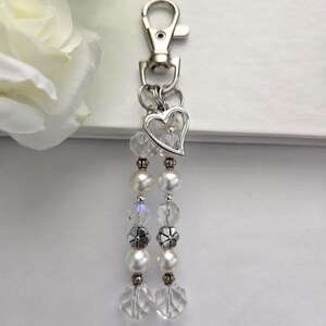 May include: A silver-toned keychain with a lobster clasp, a heart-shaped charm, and two beaded strands. The strands feature clear faceted beads, faux pearls, and silver-toned decorative beads. The keychain is designed for keys or as a decorative accessory.