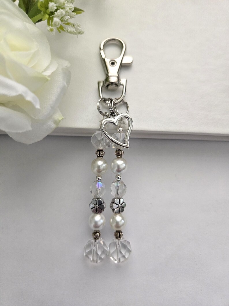 May include: A silver-toned keychain with a lobster clasp, a heart-shaped charm, and two beaded strands. The strands feature clear faceted beads, faux pearls, and silver-toned decorative beads. The keychain is designed for keys or as a decorative accessory.
