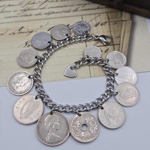 K&ouml;nnte beinhalten: Ein silberfarbenes Charm-Armband mit Kettendesign, verziert mit verschiedenen M&uuml;nz-Anh&auml;ngern. Das Armband enth&auml;lt einen herzf&ouml;rmigen Anh&auml;nger und einen Verschluss. Die M&uuml;nzen sind in verschiedenen Gr&ouml;&szlig;en und Designs.