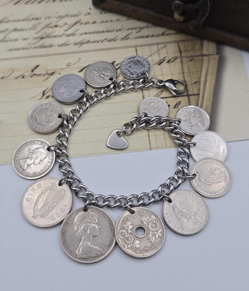 K&ouml;nnte beinhalten: Ein silberfarbenes Charm-Armband mit Kettendesign, verziert mit verschiedenen M&uuml;nz-Anh&auml;ngern. Das Armband enth&auml;lt einen herzf&ouml;rmigen Anh&auml;nger und einen Verschluss. Die M&uuml;nzen sind in verschiedenen Gr&ouml;&szlig;en und Designs.