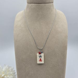 May include: A silver-toned chain necklace featuring a rectangular off-white mahjong tile pendant. The tile displays the black Chinese character for six and the red character for ten thousand. A small red bead and a tiny blue bead accent the top.